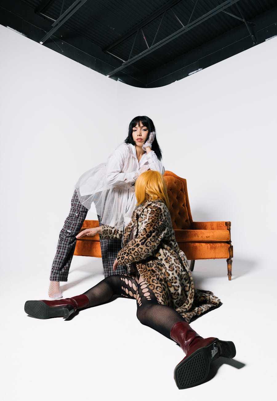 Two women in stylish outfits pose in a modern studio setting with vibrant armchair.