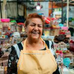 Smiling woman vendor at a lively indoor market surrounded by colorful merchandise.