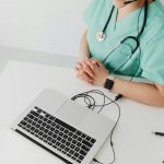Doctor in scrubs with stethoscope conducting online consultation using a laptop.