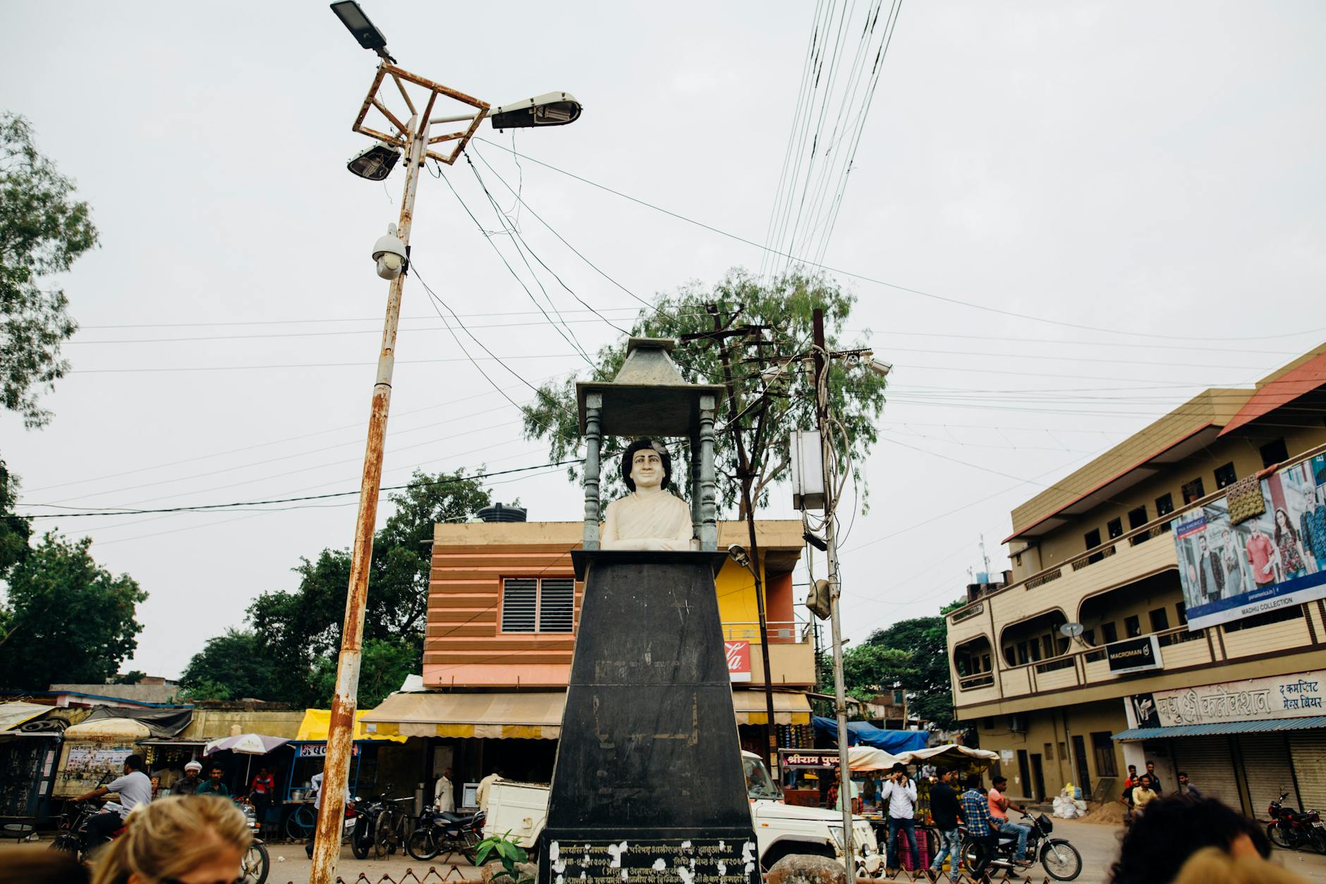 Street view of a bust sculpture in an Indian town square, surrounded by buildings and people.