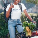Smiling African American woman on bicycle delivering groceries outdoors, showing thumbs up.