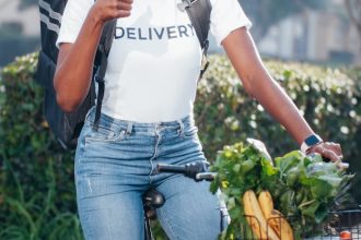 Smiling African American woman on bicycle delivering groceries outdoors, showing thumbs up.
