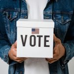 Person in denim holding a ballot box with a US flag, emphasizing voting rights.