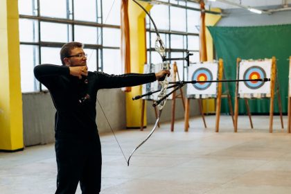 A man practicing archery indoors, aiming at a bullseye target with focused precision.