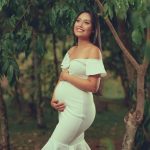 Smiling pregnant woman in a white dress, embracing her belly in a verdant outdoor setting.