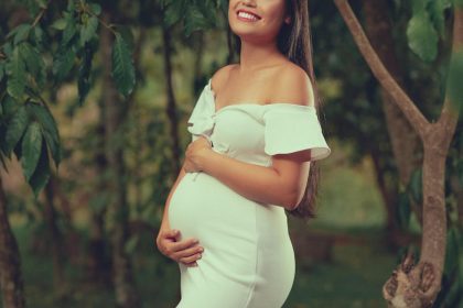 Smiling pregnant woman in a white dress, embracing her belly in a verdant outdoor setting.