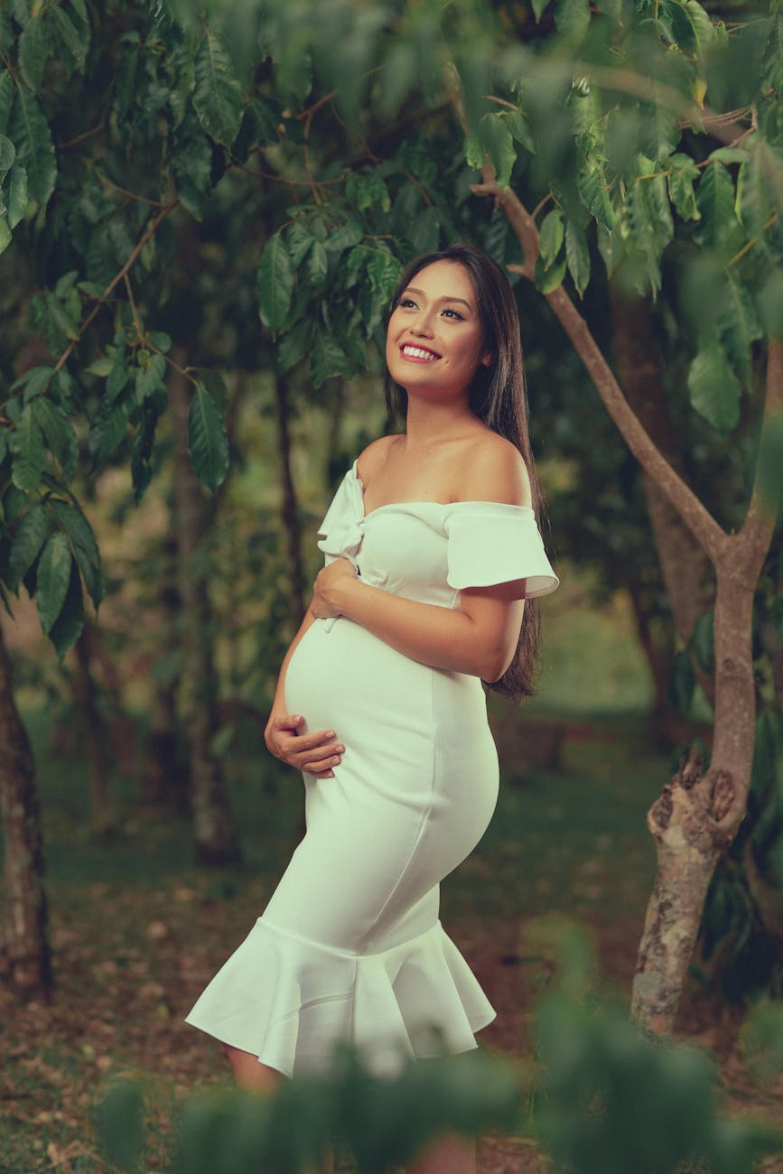 Smiling pregnant woman in a white dress, embracing her belly in a verdant outdoor setting.