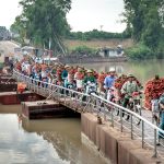 Motorbike vendors carrying fruit baskets across a rural footbridge over a river.