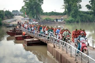 Motorbike vendors carrying fruit baskets across a rural footbridge over a river.