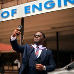 Proud graduate taking a selfie outside engineering building with sign.