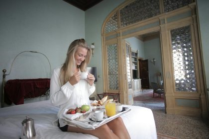 Smiling woman enjoys breakfast in a comfortable, luxurious bedroom setting.