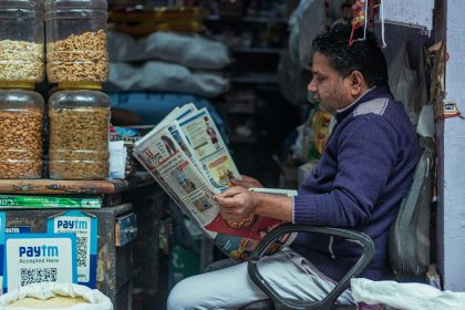 Man reading a newspaper in a small shop, surrounded by jars of snacks. Indoor market scene.