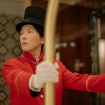 An attentive bellboy in a classic red uniform assists guests by pushing a luggage cart in a hotel lobby.