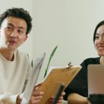 Two colleagues brainstorming at a workplace with laptops, papers, and smiles.